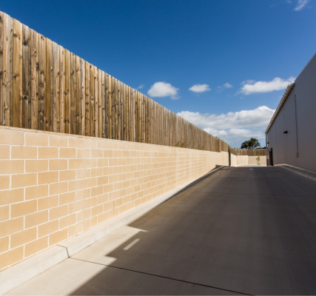 Smooth_cream_coloured_block_retaining_wall_with_white_mortar_topped_with_a_timber_fence_used_beside_the_driveway_of_an_industrial_building