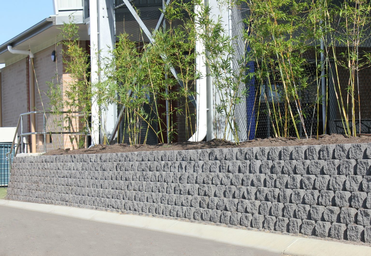 High_drystack_charcoal_retaining_wall_supporting_a_garden_bed_in_front_of_tennis_courts_at_a_school